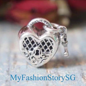 May include: A silver heart-shaped charm featuring a lattice pattern and a central keyhole design. The top of the charm is engraved with the text LOVE YOU and includes a small attached key. The piece has a polished metallic finish with dark accents.