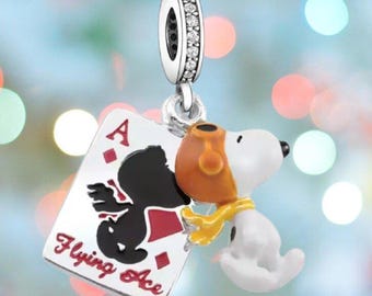 May include: A silver charm featuring Snoopy as the Flying Ace, with a playing card that reads "Flying Ace". Snoopy is wearing a brown aviator helmet and a yellow scarf. The charm has a loop with clear stones.