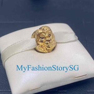 Puede incluir: Un charm dorado con forma de cabeza de león, apoyado sobre un cojín blanco con una cinta color crema. El charm tiene rasgos faciales detallados. El texto "MyFashionStorySG" está impreso en el cojín.