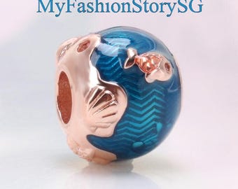 Puede incluir: Un charm de oro rosa con un diseño de globo esmaltado azul, con detalles de peces y conchas marinas. El charm tiene forma redonda y un agujero para enhebrar en una pulsera o collar. El texto "MyFashionStorySG" está en el fondo.