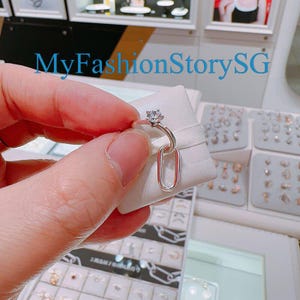 Puede incluir: Un anillo de plata con una pequeña piedra transparente y un colgante ovalado. El anillo está en una tarjeta de presentación blanca.