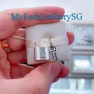 May include: A silver charm set featuring a padlock with a keyhole and a small key charm with the word "LOVE" engraved on it. The charms are displayed on a white satin background.