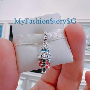 May include: A silver charm in the form of a nutcracker soldier, featuring a blue crown and red and green details, with a twisted top. The charm is displayed on a white pillow with a ribbon. The image includes the text "MyFashionStorySG".