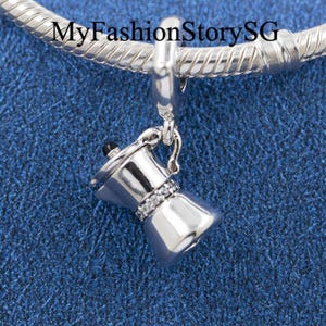 Puede incluir: Un charm plateado con forma de cafetera moka, que presenta un pequeño pomo negro en la tapa y una banda de piedras transparentes alrededor del centro. El charm cuelga de una pulsera de cadena de serpiente plateada. El texto MyFashionStorySG es visible.