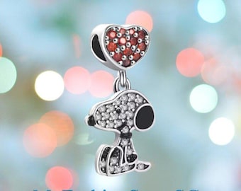May include: A silver charm featuring Snoopy with a heart-shaped top. The heart is filled with red stones, and Snoopy is adorned with clear stones and black accents. The charm is suspended from a silver loop. The text "MyFashionStorySG" is at the bottom.