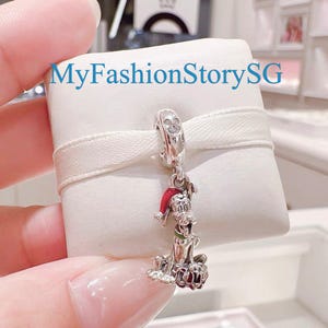 Könnte beinhalten: Ein silberner Charm-Anhänger mit einem Cartoon-Hund, der einen roten Weihnachtsmütze und einen grünen Kragen trägt. Der Anhänger hängt an einer Silberschlaufe mit einem klaren Edelstein. Der Text "MyFashionStorySG" ist sichtbar.