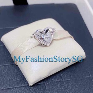 Puede incluir: Un charm de plata en forma de corazón con alas. El corazón tiene la palabra "Mom" inscrita. El charm se muestra en una caja blanca con una cinta rosa.