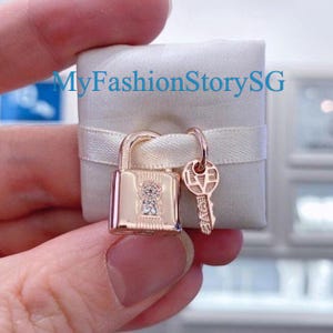 May include: A rose gold padlock charm with a key, resting on a white satin-wrapped square. The padlock has a keyhole design with a small gem. The key has the word "LOVE" engraved on it. The text "MyFashionStorySG" is visible.