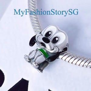 Könnte beinhalten: Silberner Anhänger mit einem Cartoon-Hund mit schwarzen Ohren, einer schwarzen Nase und einem grünen Halsband. Der Hund sitzt mit gekreuzten Pfoten und hat einen freundlichen Ausdruck.