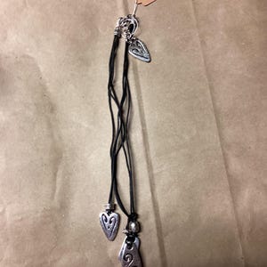 May include: A set of three silver heart-shaped pendants on black leather cords. The pendants have intricate designs and are tied together with a silver bead.