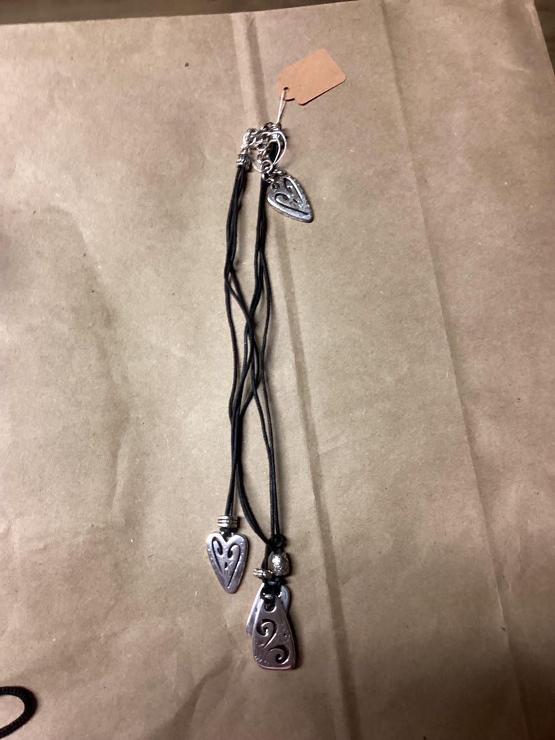May include: A set of three silver heart-shaped pendants on black leather cords. The pendants have intricate designs and are tied together with a silver bead.
