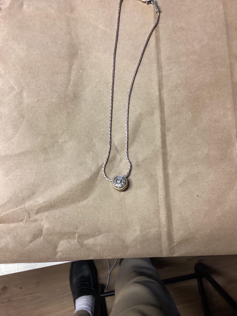 May include: A silver chain necklace with a round pendant featuring a detailed design. The pendant is gold-toned and has a raised centre.