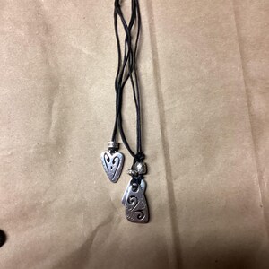 May include: A black leather cord necklace with three silver pendant charms. The charms are shaped like hearts and have intricate designs.