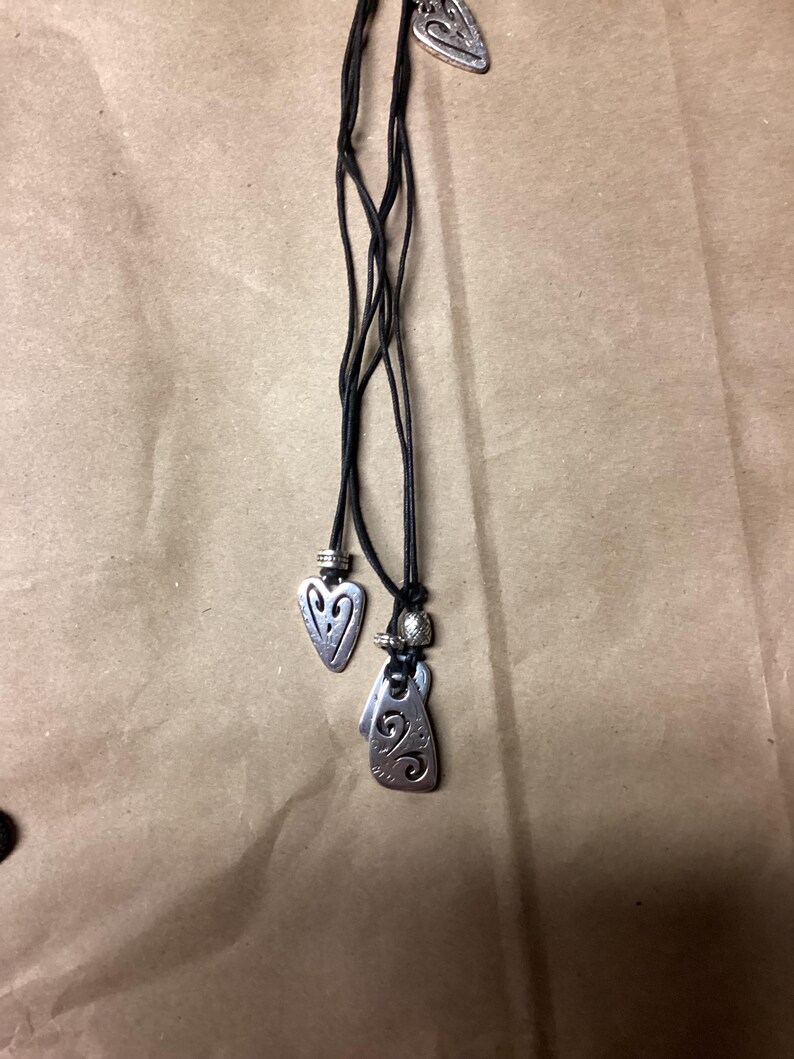May include: A black leather cord necklace with three silver pendant charms. The charms are shaped like hearts and have intricate designs.
