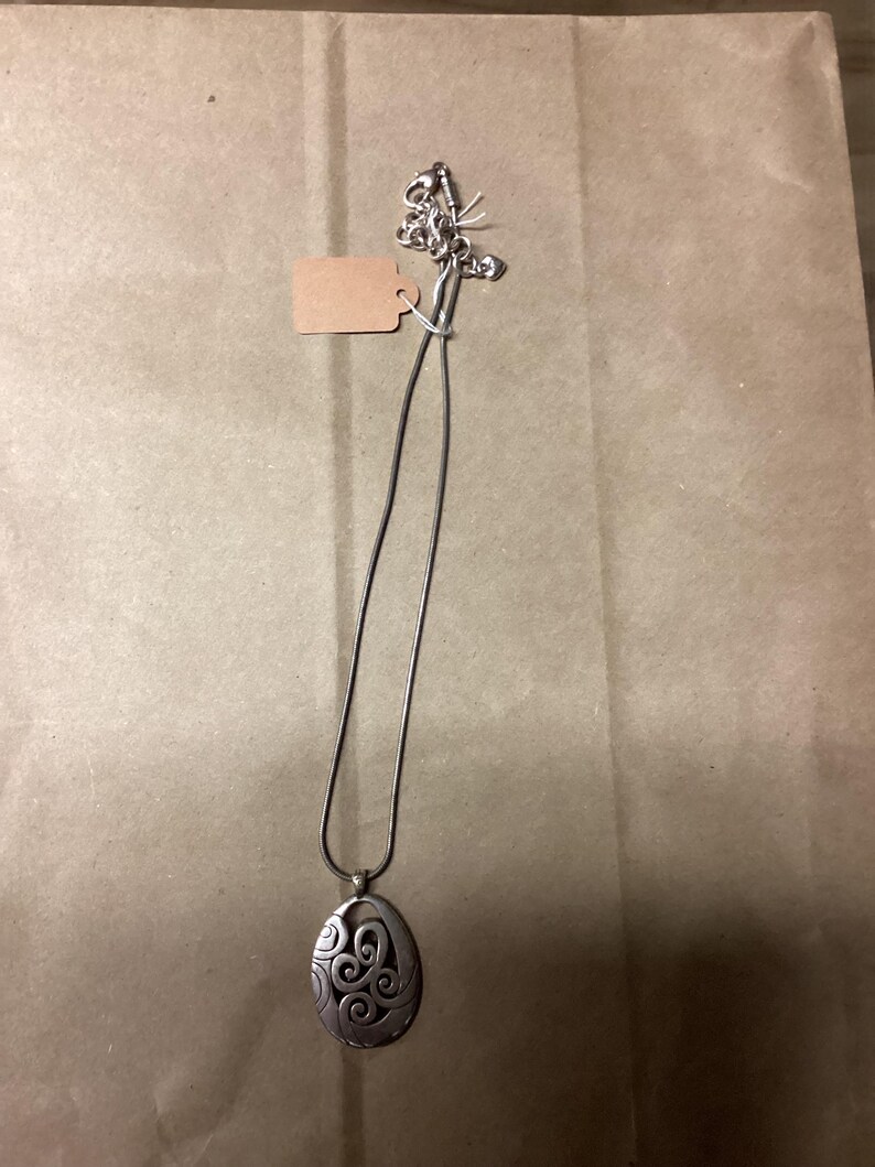 Puede incluir: Un collar de plata con un colgante con un dise&ntilde;o de espiral. El colgante es de forma ovalada y cuelga de una fina cadena de plata.