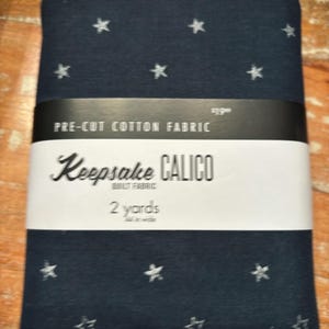 May include: A package of pre-cut cotton fabric with a navy blue background and white stars. The fabric is labeled "Keepsake Calico Quilt Fabric" and "2 yards".