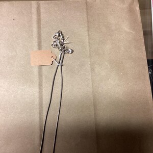 Puede incluir: Un collar de cadena de plata con un peque&ntilde;o dije. La cadena est&aacute; sobre una superficie marr&oacute;n.