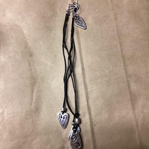 May include: A black leather cord necklace with three silver pendant charms. The charms are shaped like hearts and have intricate designs.