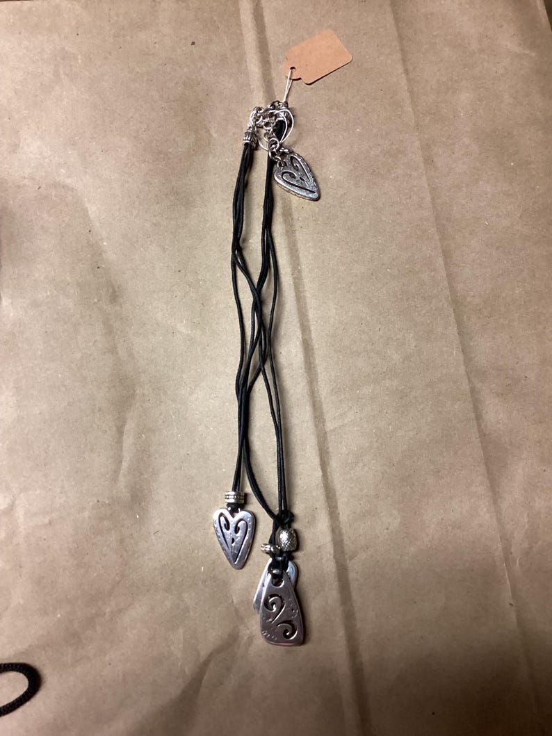 May include: A black leather cord necklace with three silver pendant charms. The charms are shaped like hearts and have intricate designs.