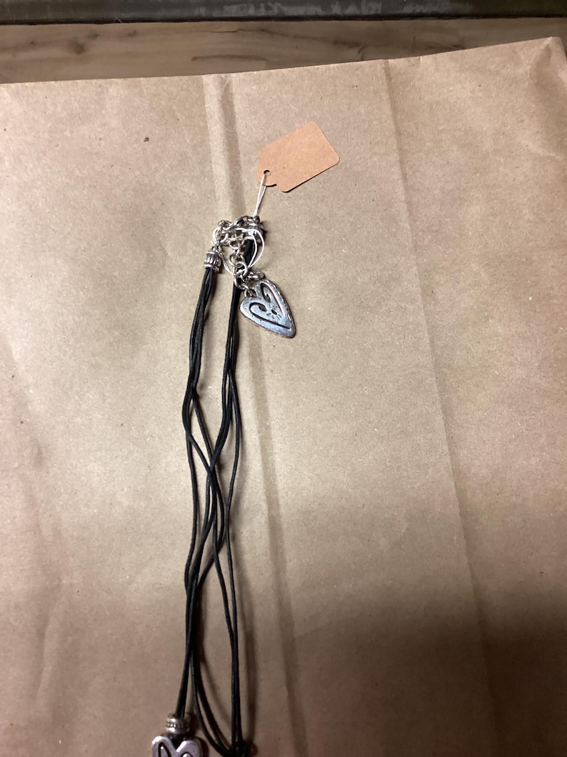 May include: A black leather cord necklace with two silver heart charms. The necklace is on a brown paper background.