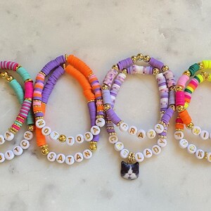 May include: Four colorful beaded bracelets with white letter beads spelling out phrases like "Karma's a Bitch" and "Not Today Satan." One bracelet has a cat charm. The bracelets feature gold accents and a variety of bright colors.