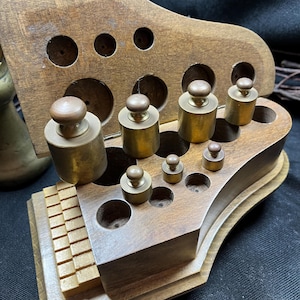 May include: A wooden scale weight set featuring a hinged box with multiple circular cutouts for storage. The set includes several cylindrical brass weights of varying sizes, each topped with a small knob, arranged within the wooden base.