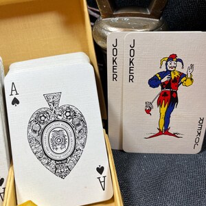 May include: A deck of playing cards in a gold box. The Ace of Spades is visible, featuring an ornate design. Two Joker cards are also visible, with a colorful jester illustration. The cards are in a gold box.