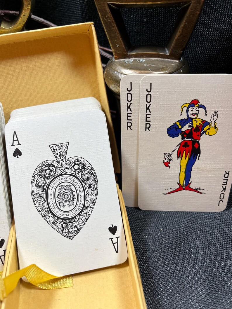 May include: A deck of playing cards in a gold box. The Ace of Spades is visible, featuring an ornate design. Two Joker cards are also visible, with a colorful jester illustration. The cards are in a gold box.
