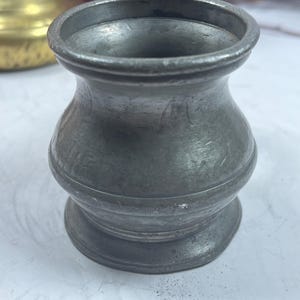 Puede incluir: Una peque&ntilde;a jarra de peltre antigua con asa. La jarra tiene un cuerpo bulboso y un borde acampanado. La superficie muestra signos de edad y desgaste, con un aspecto ligeramente deslustrado. La jarra probablemente se usa para beber.