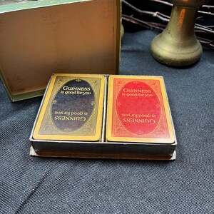 May include: A vintage card game set featuring two decks of cards with the text "Guinness is good for you" on each card. One deck has a black background, the other red. The cards are in a small, open green box.