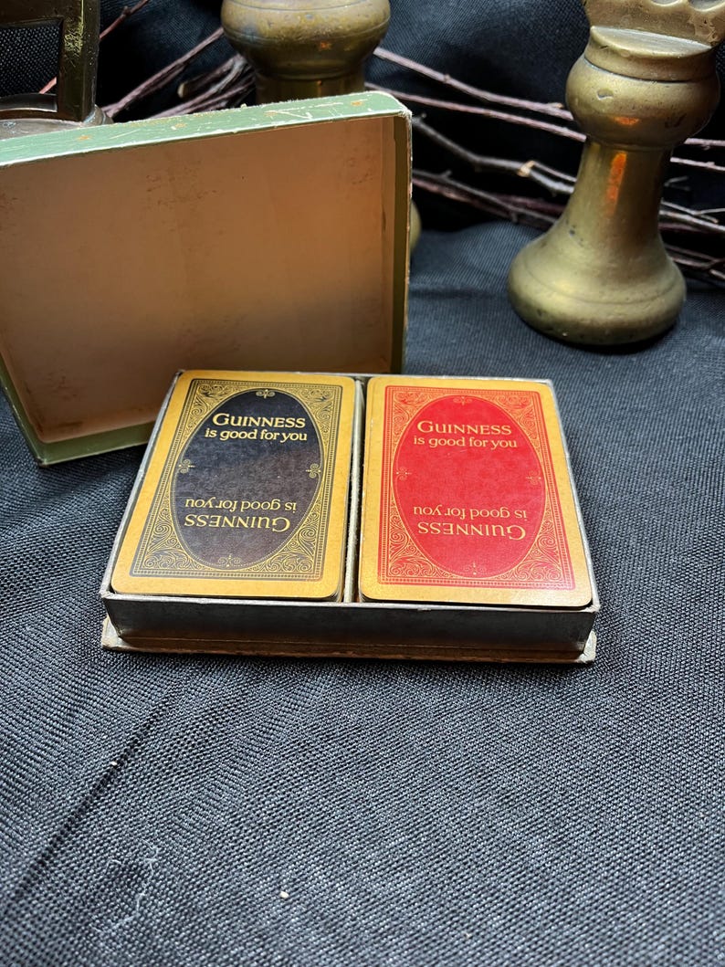 May include: A vintage card game set featuring two decks of cards with the text "Guinness is good for you" on each card. One deck has a black background, the other red. The cards are in a small, open green box.