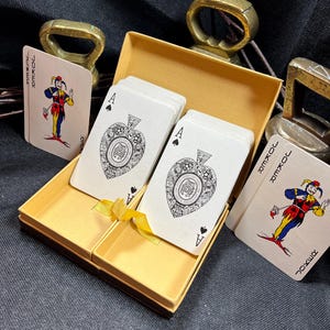 May include: A deck of playing cards in a gold-colored box. The cards feature the Ace of Spades and Joker designs. The box is open, revealing the cards inside. Two brass-colored bottle openers are in the background.