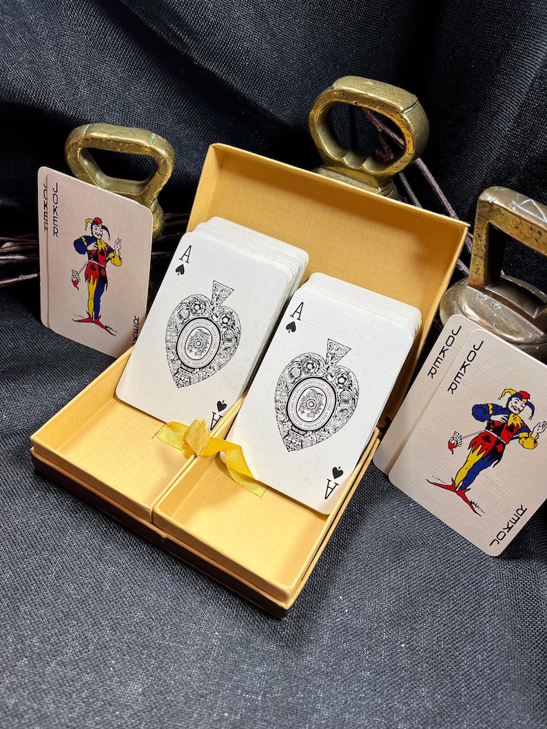 May include: A deck of playing cards in a gold-colored box. The cards feature the Ace of Spades and Joker designs. The box is open, revealing the cards inside. Two brass-colored bottle openers are in the background.