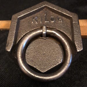 May include: A dark grey cast iron weight with a large metal ring. The word "KILOG" is embossed on the top. The ring is attached to the centre of the weight.