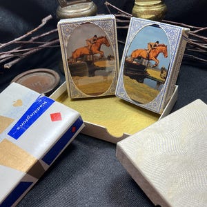 May include: Several vintage playing card decks and accessories are displayed. Two decks feature a painting of a rider on a horse jumping over a fence. One deck is labeled "Waddington's" with a blue and gold design. Other items include a brass bottle opener.