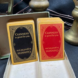 May include: Two decks of vintage Guinness playing cards. One deck has black card backs with gold text and ornate borders, the other has red card backs. The text on the cards reads "GUINNESS is good for you".
