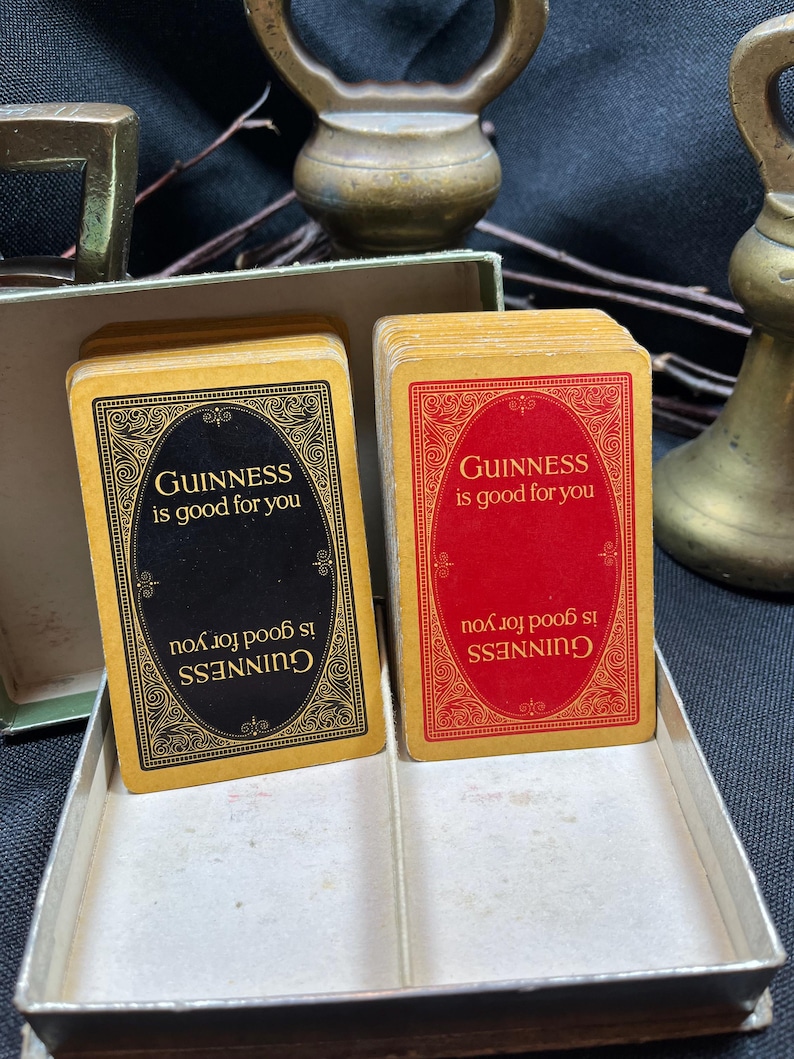 May include: Two decks of vintage Guinness playing cards. One deck has black card backs with gold text and ornate borders, the other has red card backs. The text on the cards reads "GUINNESS is good for you".