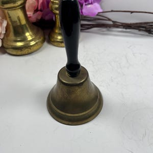 May include: A brass bell with a black wooden handle. The bell is sitting on a white surface.