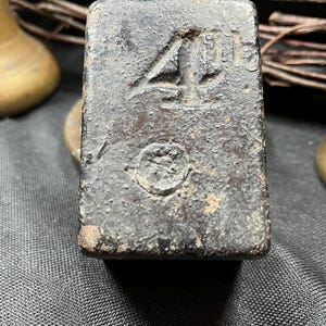 May include: A dark grey, rectangular metal weight, with the number "4" embossed on its surface. The weight has a textured appearance and a small circular marking below the number. The item appears to be vintage.