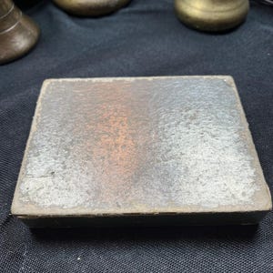 May include: A rectangular, antique box with a distressed, silver-toned surface. The box has a dark, possibly black, border. The background includes several brass-colored decorative objects.
