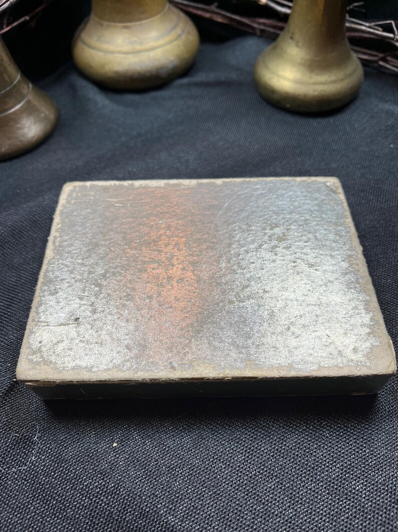 May include: A rectangular, antique box with a distressed, silver-toned surface. The box has a dark, possibly black, border. The background includes several brass-colored decorative objects.