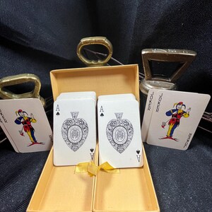 May include: A deck of playing cards in a yellow box, featuring the Ace of Spades with an ornate design. Two Jokers are visible, along with two brass bottle openers. The cards are displayed on a dark background.