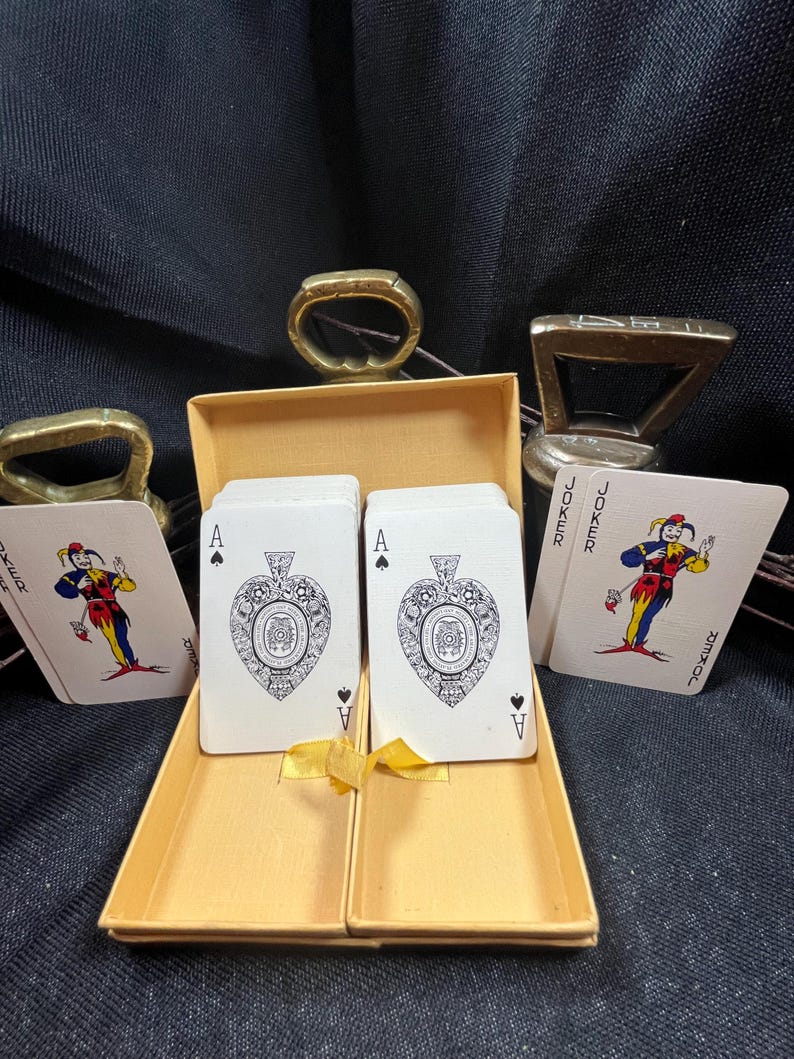 May include: A deck of playing cards in a yellow box, featuring the Ace of Spades with an ornate design. Two Jokers are visible, along with two brass bottle openers. The cards are displayed on a dark background.