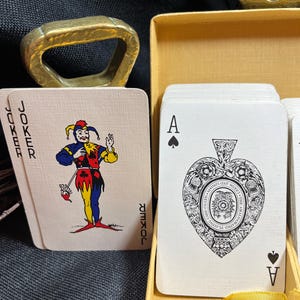 May include: A deck of playing cards with a joker card featuring a colorful jester and an Ace of Spades card with an ornate heart design. The cards are in a yellow box with a gold bottle opener.