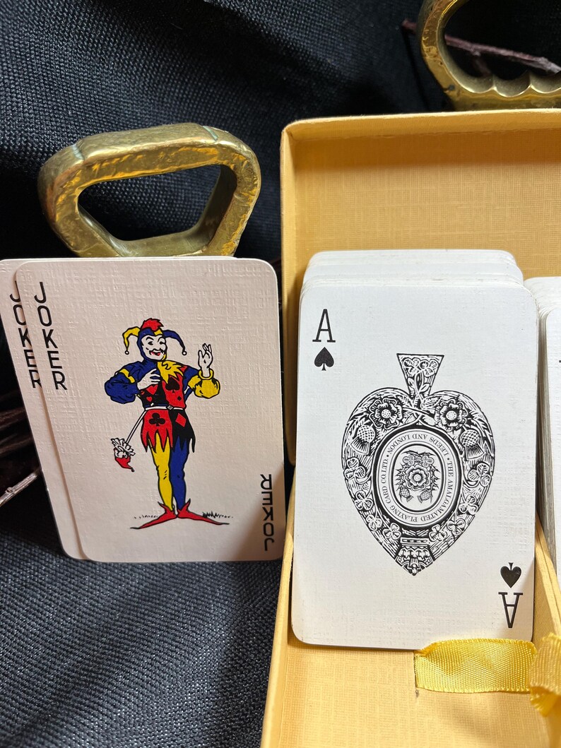 May include: A deck of playing cards with a joker card featuring a colorful jester and an Ace of Spades card with an ornate heart design. The cards are in a yellow box with a gold bottle opener.