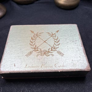 May include: A vintage playing card box with a light green and gold textured surface. The box features a gold emblem of crossed arrows and laurel leaves, with the words "Embassy Playing Cards" printed below.