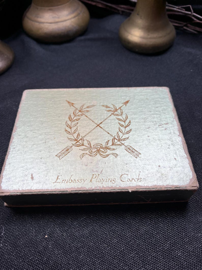 May include: A vintage playing card box with a light green and gold textured surface. The box features a gold emblem of crossed arrows and laurel leaves, with the words "Embassy Playing Cards" printed below.