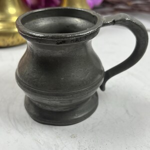 Puede incluir: Una peque&ntilde;a jarra antigua de peltre con un asa curvada. La jarra tiene un cuerpo bulboso y un borde acampanado. La palabra "HALF" est&aacute; estampada en el borde. La jarra es de color gris oscuro y parece envejecida.