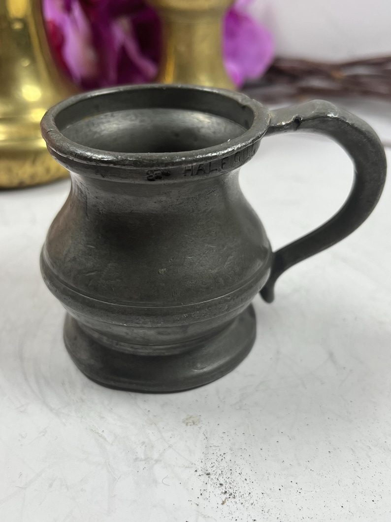 Puede incluir: Una peque&ntilde;a jarra antigua de peltre con un asa curvada. La jarra tiene un cuerpo bulboso y un borde acampanado. La palabra "HALF" est&aacute; estampada en el borde. La jarra es de color gris oscuro y parece envejecida.