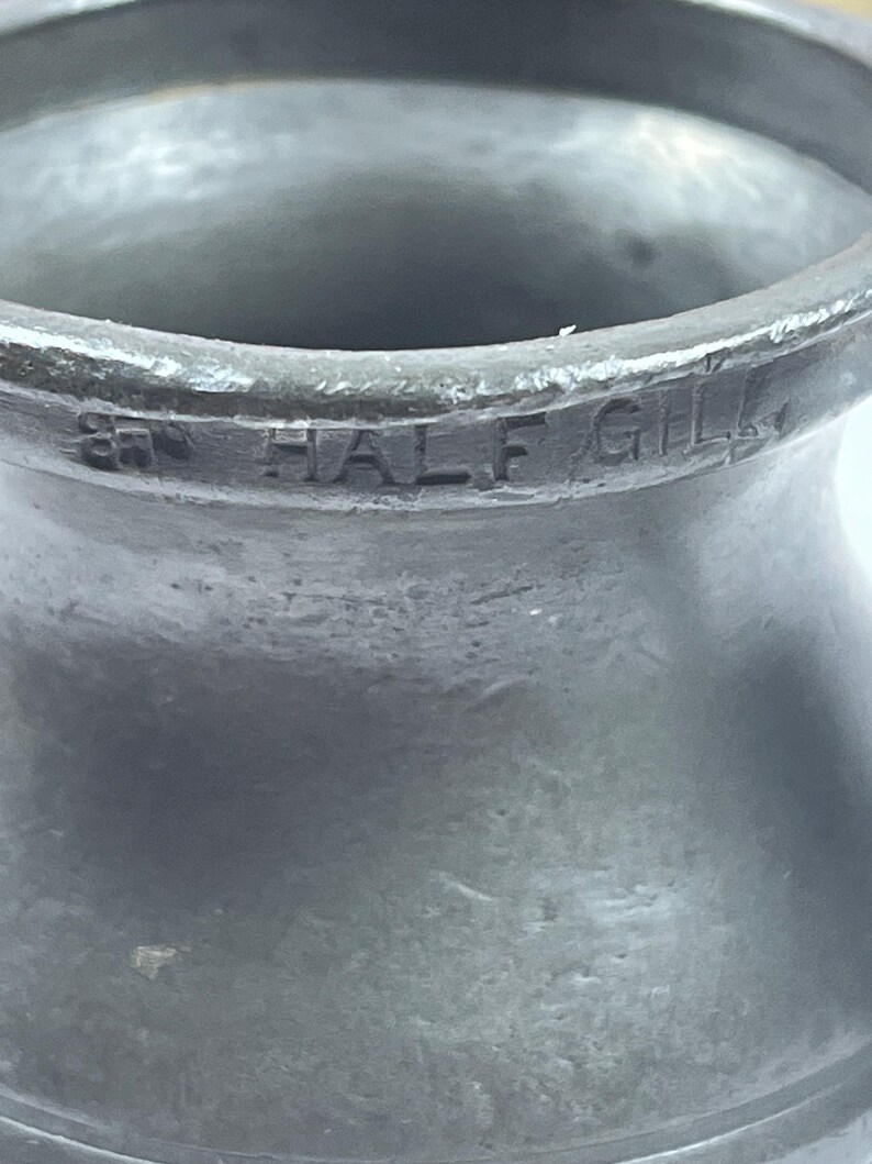 Puede incluir: Primer plano de una taza medidora vintage de metal gris. El texto "87 HALF GILL" est&aacute; grabado en el lateral. La taza tiene una superficie texturizada y un borde acampanado.